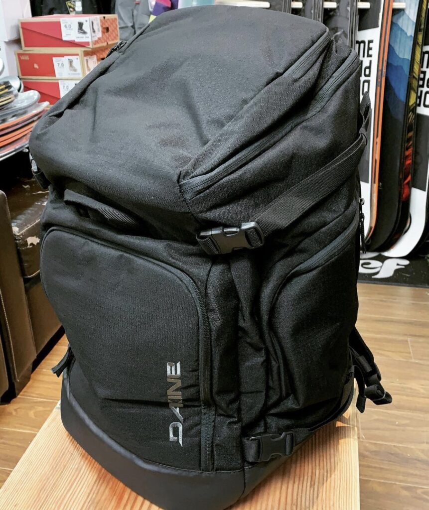 Black Dakine Boot Pack backpack standing upright in a ski shop with boards and boot boxes in the background.