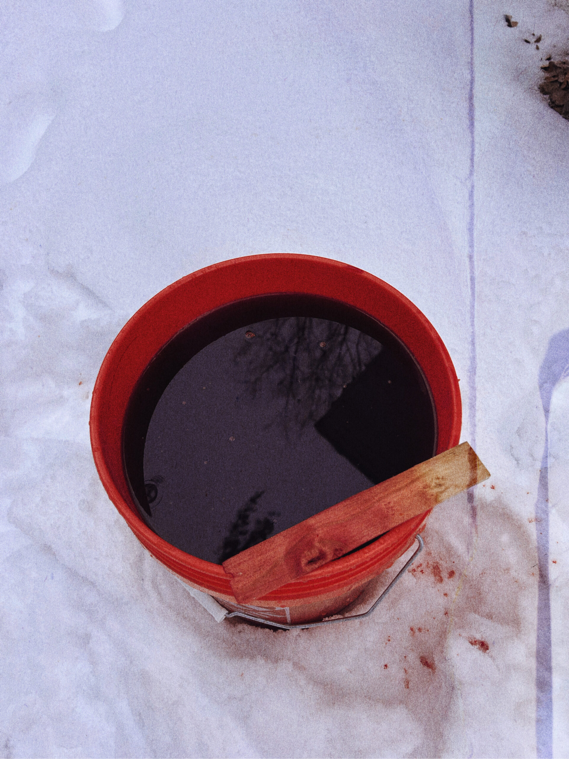 Red bucket filled with dark dye bath and a wooden stir stick resting on top