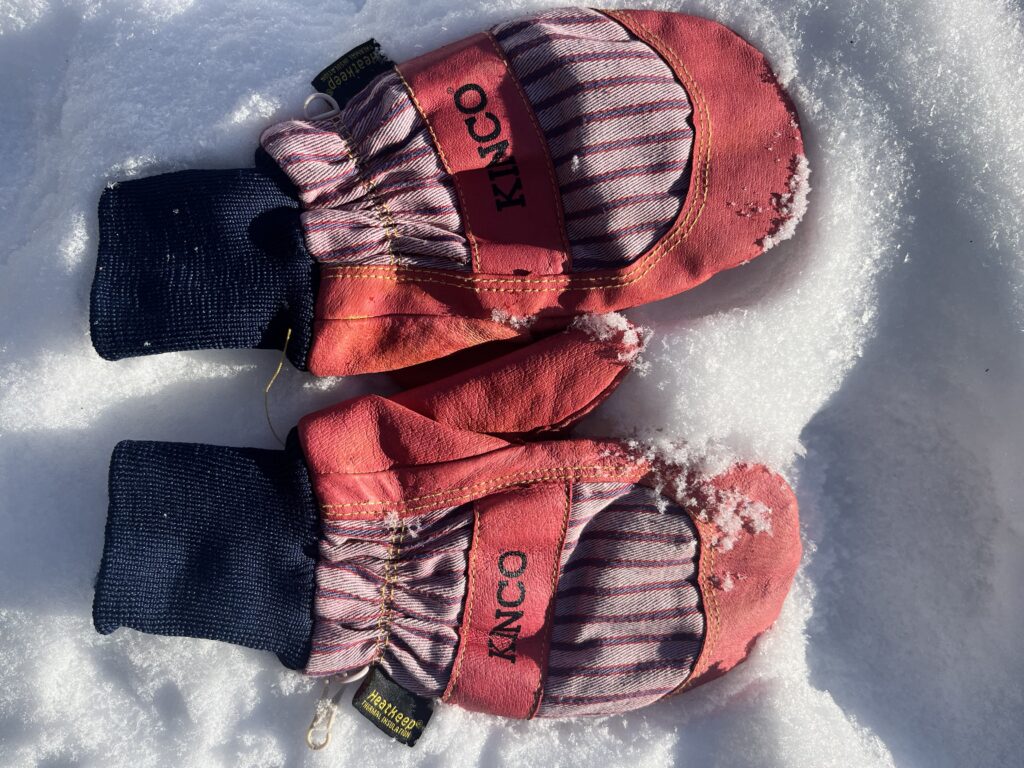 Pair of pink-dyed Kinco leather mittens with striped backs and black knit cuffs lying on fresh snow